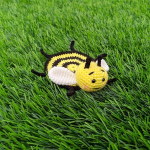 May include: A handmade crochet bee coaster with a yellow body, black stripes, and white wings. The bee has a smiling face and is lying on a bed of green artificial grass. A cute, whimsical item.