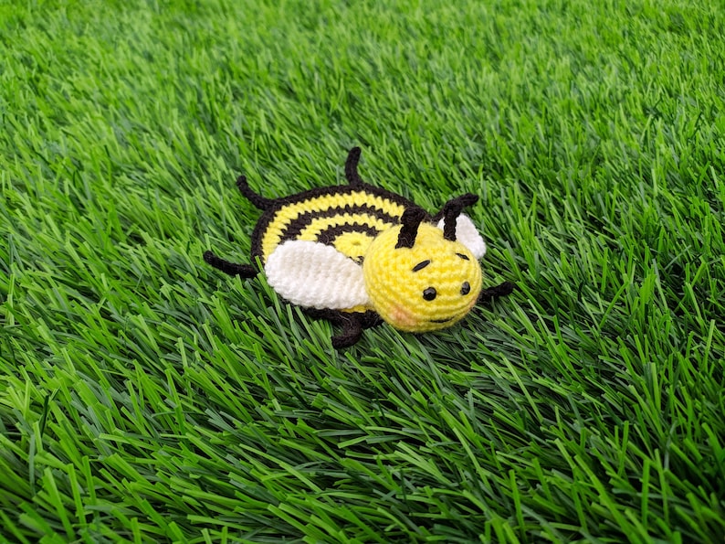 May include: A handmade crochet bee coaster with a yellow body, black stripes, and white wings. The bee has a smiling face and is lying on a bed of green artificial grass. A cute, whimsical item.