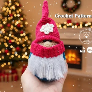May include: A small, handmade crochet gnome figure featuring a tall, pointed magenta hat adorned with a white snowflake applique, a tan nose, and a thick, textured white beard. The text "Crochet Pattern" is visible in the upper right corner.