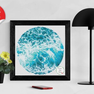 May include: A framed cross-stitch pattern of a blue and white ocean wave design. The design is circular and the text "Creative Home Patterns" is in the lower right corner.