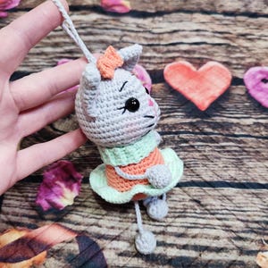 Peut inclure: Ornement de chat au crochet gris, orange et vert. Le chat porte un n&oelig;ud et une robe. L'ornement est suspendu par une ficelle grise. Le fond comprend une surface en bois et des &eacute;l&eacute;ments d&eacute;coratifs.