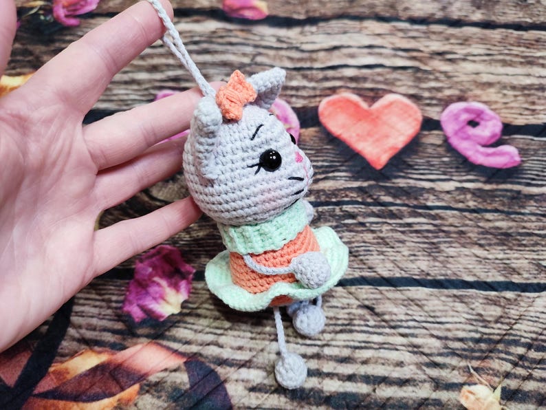 Peut inclure: Ornement de chat au crochet gris, orange et vert. Le chat porte un n&oelig;ud et une robe. L'ornement est suspendu par une ficelle grise. Le fond comprend une surface en bois et des &eacute;l&eacute;ments d&eacute;coratifs.