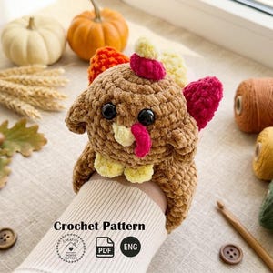 May include: A brown crocheted turkey with red, yellow, and orange accents. The turkey has large black eyes and a pink wattle. The image includes pumpkins, yarn, and the text "Crochet Pattern".