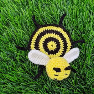 May include: A handmade crochet bee coaster with a yellow head and body, black and yellow striped abdomen, white wings, and black antennae. The bee is lying on a bed of green artificial grass.