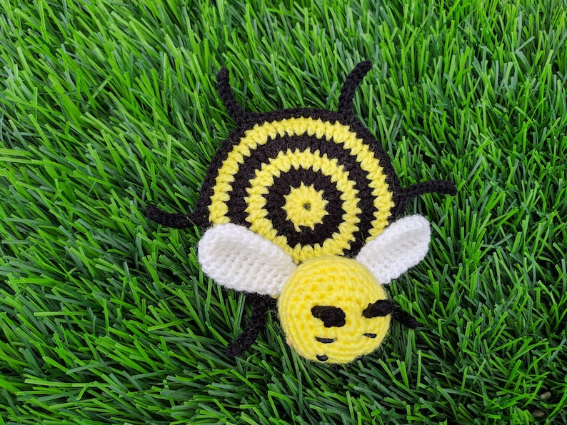 May include: A handmade crochet bee coaster with a yellow head and body, black and yellow striped abdomen, white wings, and black antennae. The bee is lying on a bed of green artificial grass.