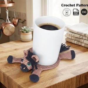 May include: A white ceramic mug filled with coffee rests on a handmade crochet horse coaster. The coaster is primarily brown with black accents and green eyes. The image also includes the text "Crochet Pattern".
