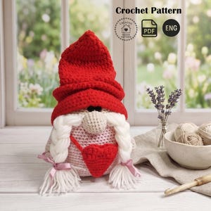 May include: A handmade crochet gnome with a red hat, white braids, and a red heart. The gnome is wearing a pink and gray body. The image includes the text "Crochet Pattern" and a PDF and ENG icon.