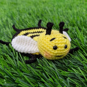 May include: A handmade crochet bee coaster toy with a yellow body, black stripes, and white wings. The bee has black eyes and antennae. The toy is lying on a bed of green artificial grass.