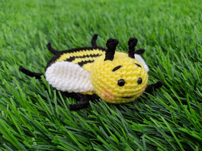 May include: A handmade crochet bee coaster toy with a yellow body, black stripes, and white wings. The bee has black eyes and antennae. The toy is lying on a bed of green artificial grass.