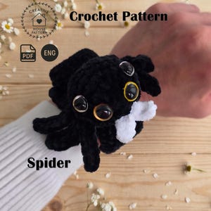 Könnte beinhalten: Eine schwarze Häkelspinne mit großen glänzenden Augen und einem weißen Akzent. Der Text "Crochet Pattern", "PDF", "ENG" und "Spider" ist sichtbar. Die Spinne ist an einem weißen gerippten Ärmel befestigt und zeigt ihre weiche Textur sowie die detaillierte Garnkonstruktion.