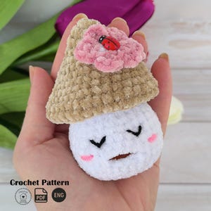 May include: A handmade crochet mushroom with a tan cap, white body, and a smiling face. A pink flower with a red ladybug sits atop the cap. The mushroom is a soft, plush toy.