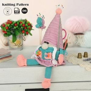 Puede incluir: Una muñeca gnomo hecha a mano con un gorro rosa tejido y un cuerpo azul, que sostiene agujas de tejer y otros suministros de artesanía. La muñeca tiene barba blanca y pies rosas. La imagen incluye el texto "Knitting Pattern" y un logotipo.