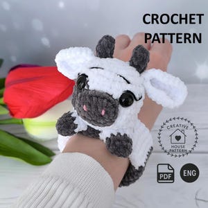 May include: A white and gray crochet cow with black eyes and pink nose. The crochet cow is being worn on a wrist. The image includes the text "CROCHET PATTERN" and a logo with the words "CREATIVE HOUSE PATTERN".