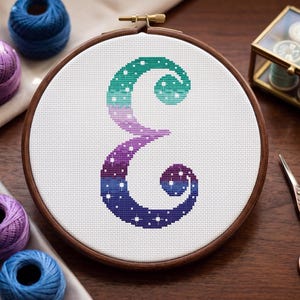 May include: Cross-stitch embroidery of the letter "E" in a wooden hoop. The letter features a gradient of colours, transitioning from teal to purple, with white star-like details. Craft supplies, including thread spools and scissors, are arranged around the hoop.