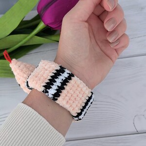 May include: A pink crochet pattern snake slap bracelet with black and white checkered accents. The snake has a red tongue and a black eye.