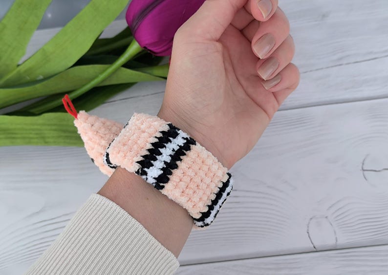 May include: A pink crochet pattern snake slap bracelet with black and white checkered accents. The snake has a red tongue and a black eye.