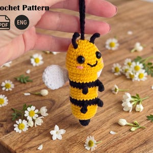 May include: A handmade crochet bee with yellow and black stripes, a white wing, and a smiling face. The bee is suspended by a black string. The image includes the text "Crochet Pattern" and the PDF and ENG icons.