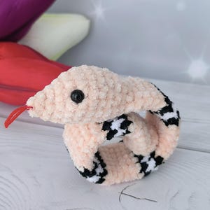 May include: A pink crochet pattern snake slap bracelet with black and white checkered accents. The snake has a red tongue and a black eye.