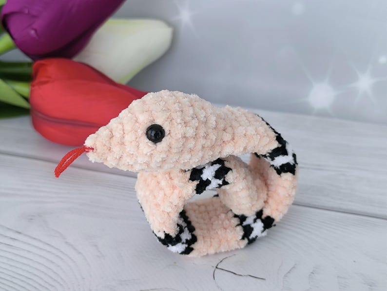 May include: A pink crochet pattern snake slap bracelet with black and white checkered accents. The snake has a red tongue and a black eye.