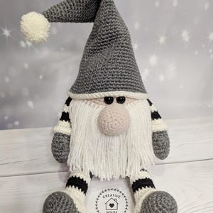 May include: A handmade crochet gnome doll with a gray pointed hat and a white pom-pom. The gnome has a white beard, a pink nose, and black eyes. The arms and legs are gray with black and white stripes. The doll is sitting on a wooden surface.