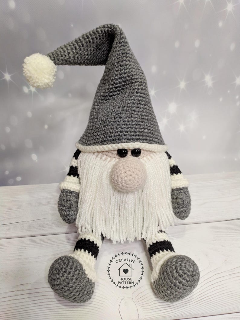 May include: A handmade crochet gnome doll with a gray pointed hat and a white pom-pom. The gnome has a white beard, a pink nose, and black eyes. The arms and legs are gray with black and white stripes. The doll is sitting on a wooden surface.
