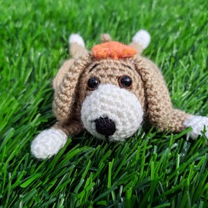 May include: A crocheted dog mug rug with a tan body, white muzzle and paws, and black eyes and nose. The dog has an orange hat and is lying on green grass.