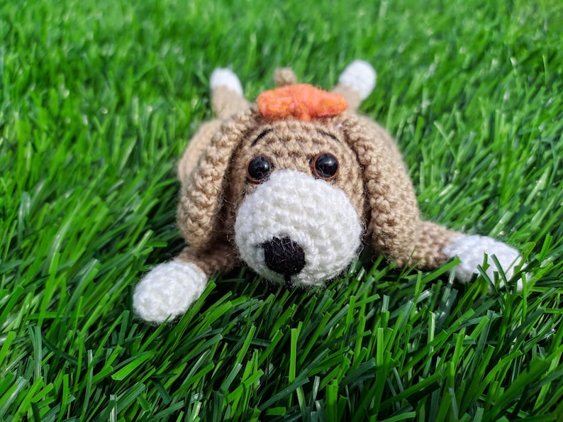 May include: A crocheted dog mug rug with a tan body, white muzzle and paws, and black eyes and nose. The dog has an orange hat and is lying on green grass.