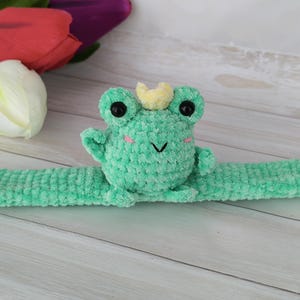 May include: A handmade, light green crochet frog with a yellow crown, black eyes, and pink cheeks. The frog is attached to a matching green slap bracelet. The background includes white, red, and purple tulips on a light wood surface.