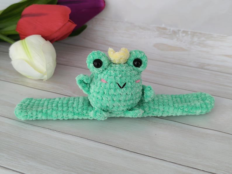 May include: A handmade, light green crochet frog with a yellow crown, black eyes, and pink cheeks. The frog is attached to a matching green slap bracelet. The background includes white, red, and purple tulips on a light wood surface.