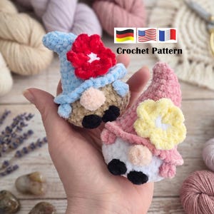 May include: Two handmade crochet gnomes with blue and pink hats, red and yellow flowers, and tan faces. The gnomes have black eyes and are held in a hand. The text "Crochet Pattern" is above the flags of Germany, the United States, and France.