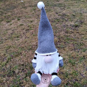 May include: A handmade crochet gnome doll with a gray pointed hat and white beard. The gnome has a pink nose, gray feet, and black and white striped arms and legs. The doll is sitting on a grassy surface.