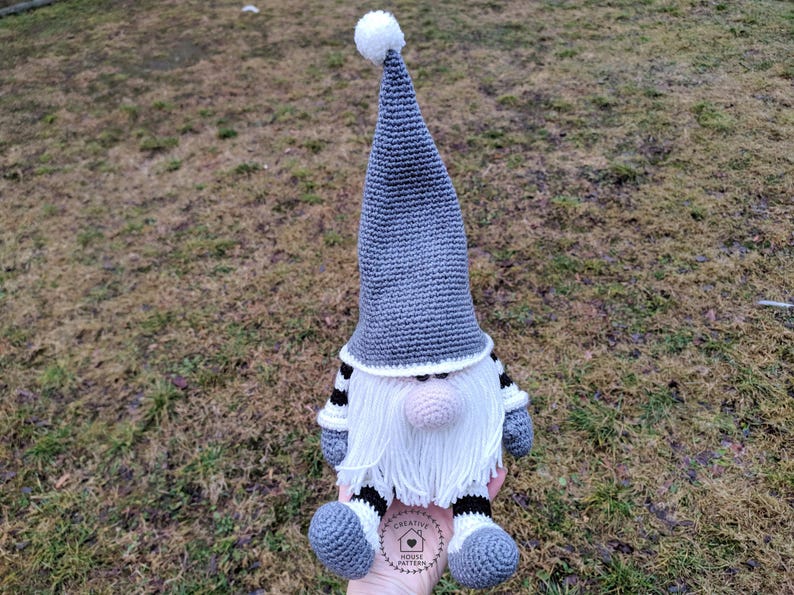 May include: A handmade crochet gnome doll with a gray pointed hat and white beard. The gnome has a pink nose, gray feet, and black and white striped arms and legs. The doll is sitting on a grassy surface.