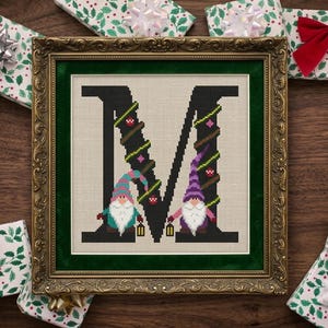 May include: Framed cross-stitch art featuring a black letter "M" decorated with Christmas lights and two gnome figures. The artwork is in a gold ornate frame with a green mat, surrounded by wrapped gifts with red bows.
