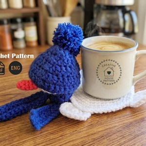 May include: A blue crochet penguin with a red beak and white belly sits next to a beige mug of coffee on a white crochet coaster. The mug has a black logo and the words "Creative House Pattern". The background includes a coffee maker and spice rack.