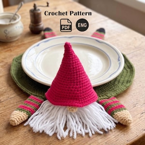 Könnte beinhalten: Ein gehäkelter Zwerg-Tischset mit roter Mütze, weißem Bart und grünem Untergrund. Das Tischset ist für einen Teller konzipiert, mit passenden armförmigen Akzenten. Der Text "Crochet Pattern" ist enthalten.
