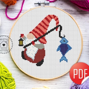 Puede incluir: Bordado de punto de cruz de un gnomo pescando, sosteniendo una linterna y usando un sombrero a rayas rojas y blancas. El gnomo sostiene una caña de pescar con un pez azul al final. El bordado está en un aro de madera.