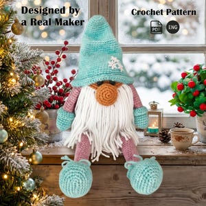 Puede incluir: Un muñeco gnomo de ganchillo con un sombrero alto azul claro con copos de nieve blancos, nariz redonda color canela, barba larga de hilo blanco, brazos y piernas rosas, y manoplas y zapatos azul claro. Texto: Designed by a Real Maker Crochet Pattern.