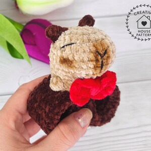May include: Sensory Capybara Fidget Toy Crochet Pattern with a red bow tie. The Capybara has a light brown face and dark brown ears and body. The toy is held in a person's hand, with a blurred background of flowers.