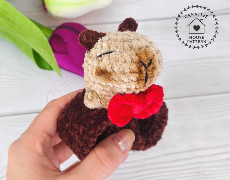 May include: Sensory Capybara Fidget Toy Crochet Pattern with a red bow tie. The Capybara has a light brown face and dark brown ears and body. The toy is held in a person's hand, with a blurred background of flowers.