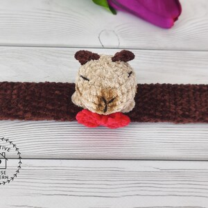 May include: A handmade brown and tan plush animal head with a red bow tie, attached to a long brown plush band. The plush animal has small brown ears and is centered on the band. The item is on a white wooden surface. Sensory Capybara Fidget Toy Crochet Pattern