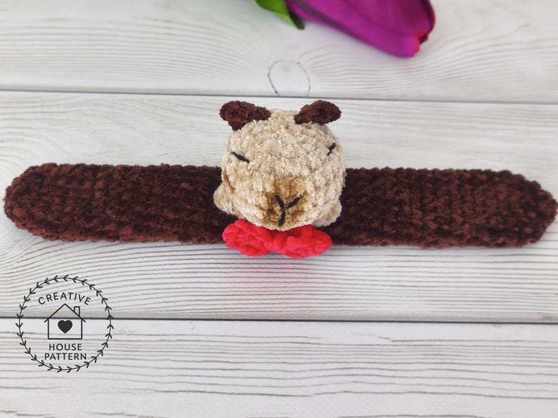 May include: A handmade brown and tan plush animal head with a red bow tie, attached to a long brown plush band. The plush animal has small brown ears and is centered on the band. The item is on a white wooden surface. Sensory Capybara Fidget Toy Crochet Pattern