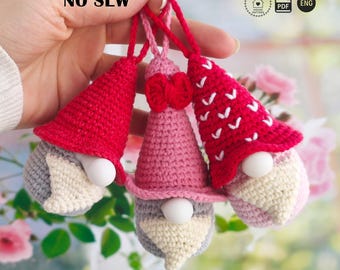 Puede incluir: Tres adornos de gnomos de ganchillo hechos a mano. Cada gnomo tiene un gorro puntiagudo rojo o rosa, una barba blanca y una nariz redonda blanca. Los gnomos se sujetan con una cuerda roja. El texto "NO SEW" es visible.