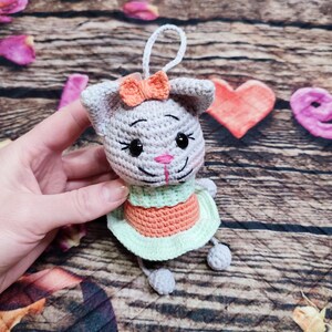 Peut inclure: Une poup&eacute;e chat au crochet faite &agrave; la main avec un corps gris, un n&oelig;ud orange et une robe verte et orange. La poup&eacute;e a des yeux noirs et un nez rose. L'arri&egrave;re-plan comprend des planches de bois et des &eacute;l&eacute;ments d&eacute;coratifs.