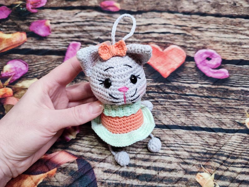 Peut inclure: Une poup&eacute;e chat au crochet faite &agrave; la main avec un corps gris, un n&oelig;ud orange et une robe verte et orange. La poup&eacute;e a des yeux noirs et un nez rose. L'arri&egrave;re-plan comprend des planches de bois et des &eacute;l&eacute;ments d&eacute;coratifs.