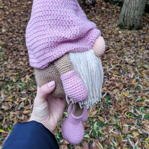 May include: A handmade crochet gnome doll with a pink hat, shoes, and mittens. The gnome has a beige body, a peach nose, and a long white beard. The doll is held in a person's hand.