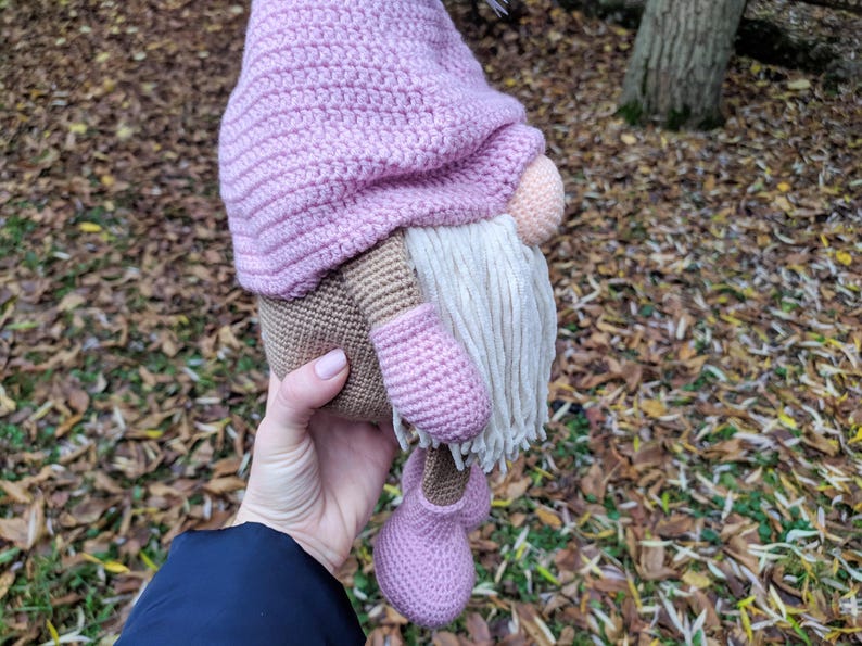 May include: A handmade crochet gnome doll with a pink hat, shoes, and mittens. The gnome has a beige body, a peach nose, and a long white beard. The doll is held in a person's hand.