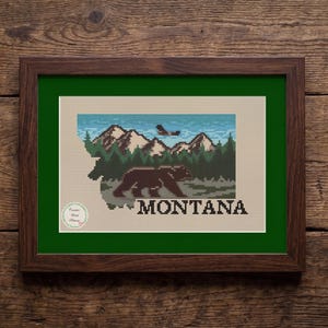 May include: A framed cross-stitch pattern shaped like the state of Montana, featuring a brown bear walking through a forest, snow-capped mountains, a flying bird, and a blue sky. The word MONTANA is stitched in black at the bottom right corner.