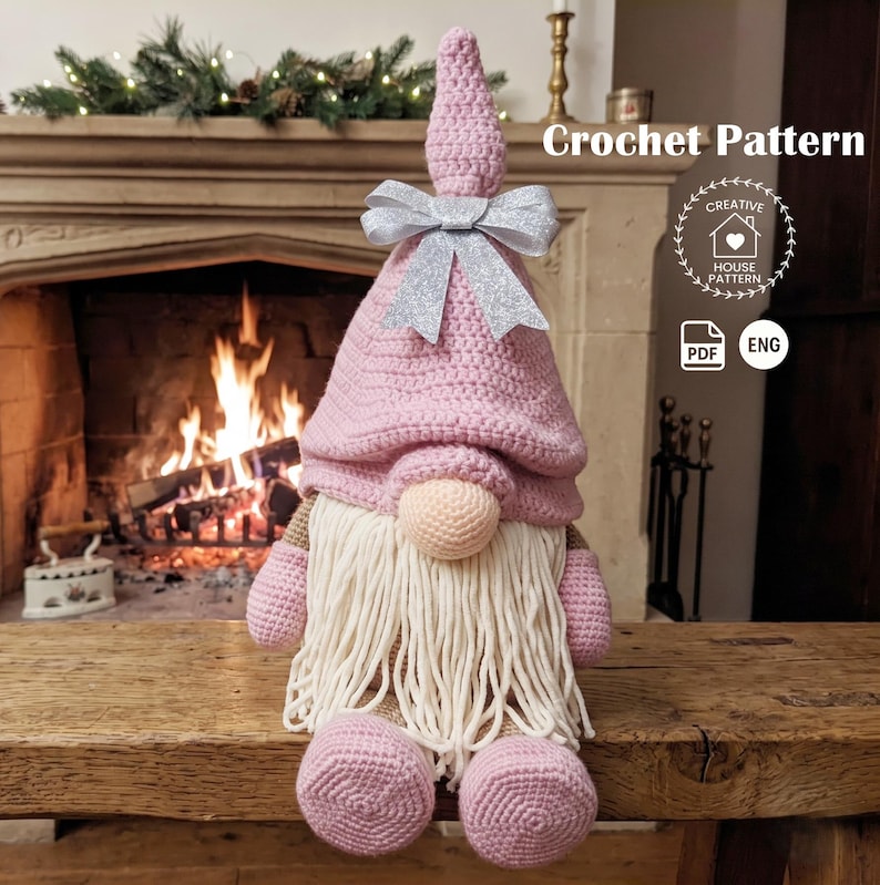 A crocheted Christmas gnome with a long white beard and a pink conical hat adorned with a shiny silver ribbon, sitting on a wooden bench in front of a cozy, lit fireplace. The image shows the final product created from the digital PDF pattern.