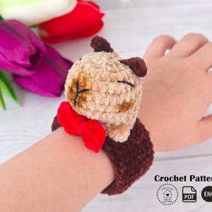 May include: A crocheted brown and tan bear wristband with a red bow tie. The bear has closed eyes and is attached to a dark brown wristband. The image also includes the text "Crochet Pattern".