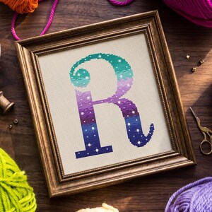 Op de afbeelding: Een ingelijste kruissteek van de letter "R" in een verloop van teal, paars en blauw, met witte steraccenten. De borduursel is geplaatst in een goudkleurige houten lijst. Garen en knutselspullen omringen de lijst.